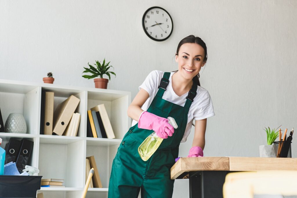 beautiful young cleaner holding spray bottle with detergent and smiling at camera while cleaning office