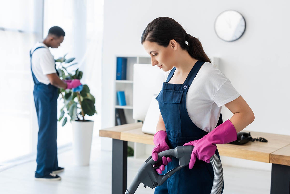 a man cleaning the plants and a woman vaccuming the office floor