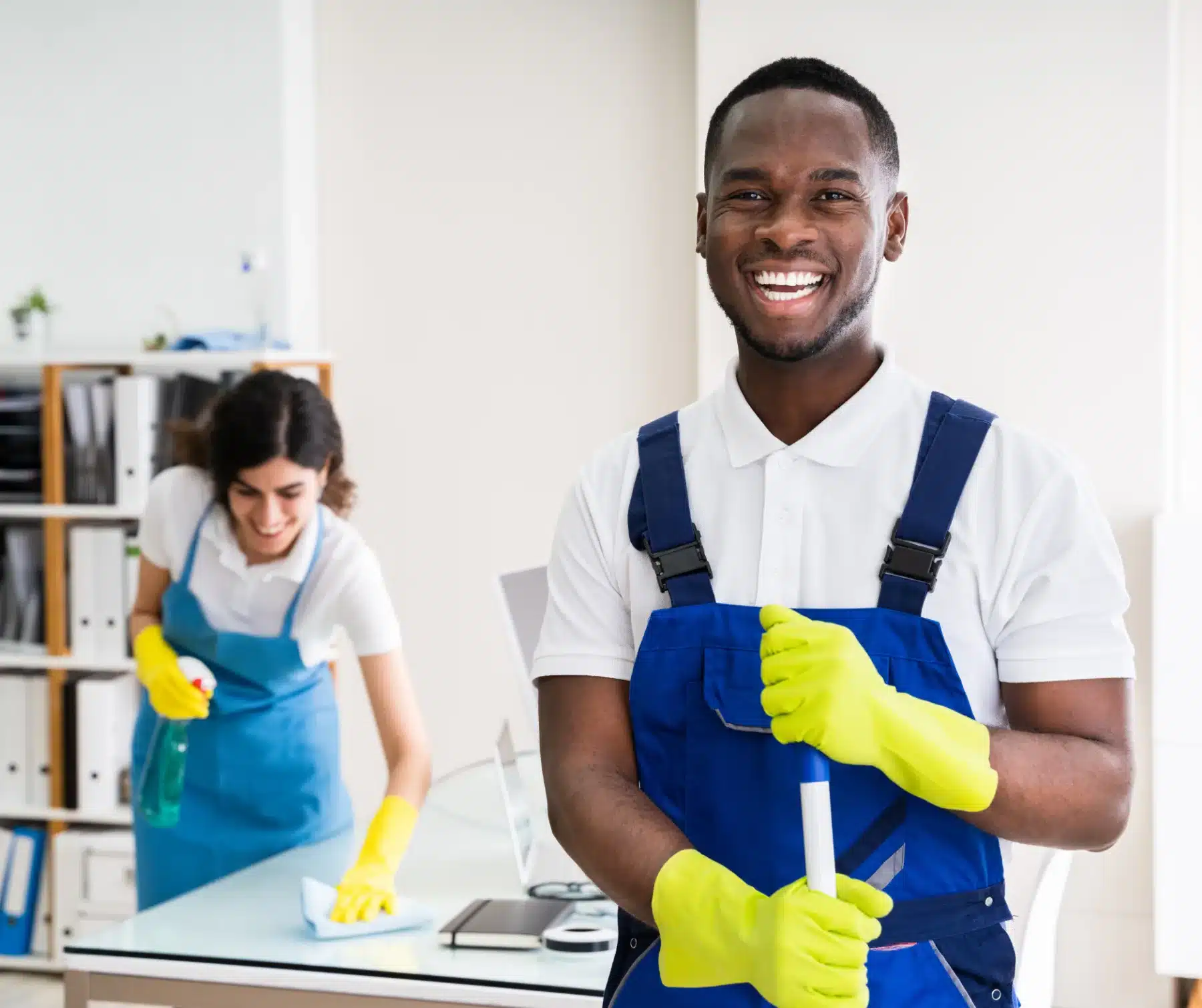 A man holding mop smiling while a woman in the back is cleaning the table